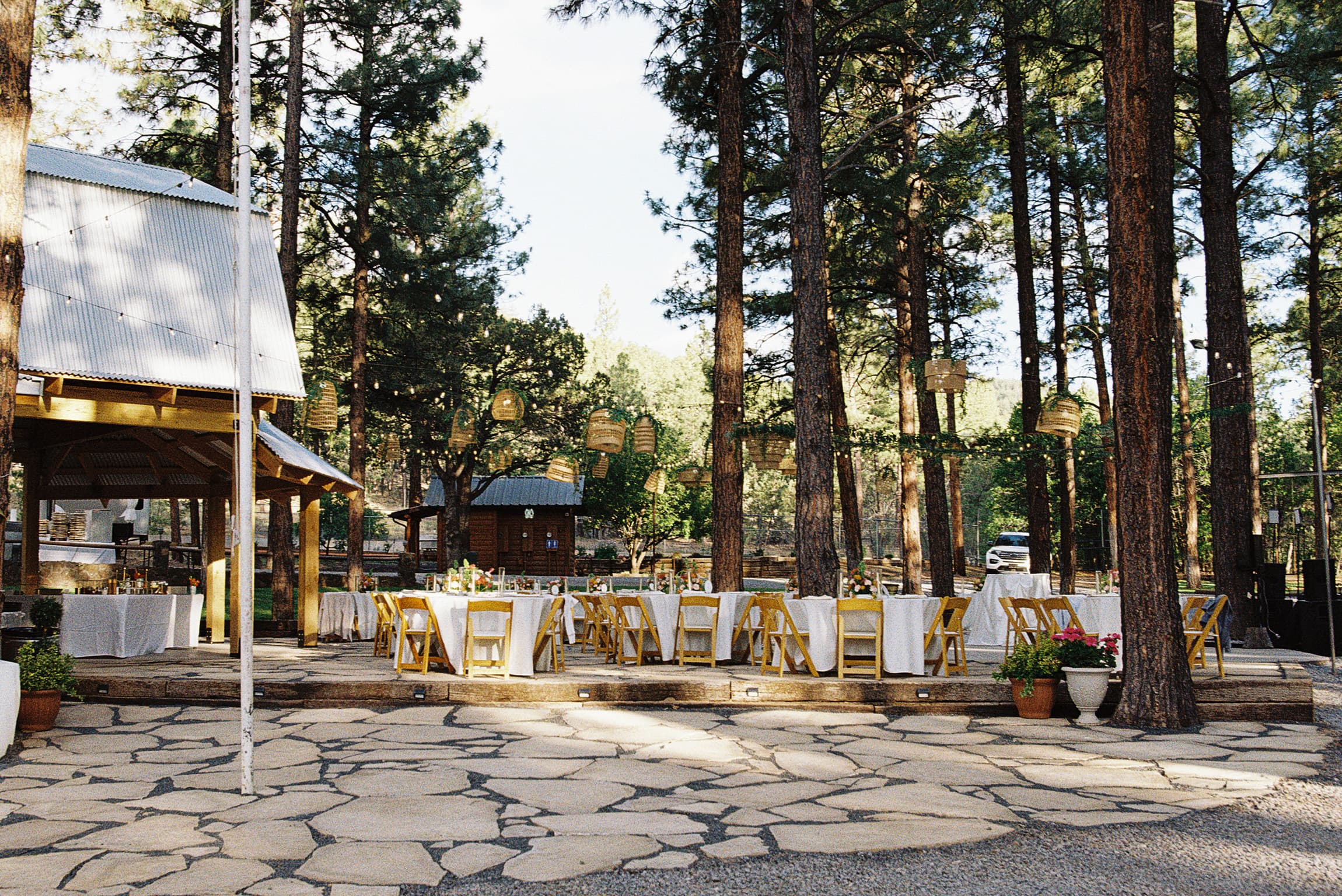 Long banquet tables set for a reception on the flagstone patio beneath towering pines and hanging lanterns
